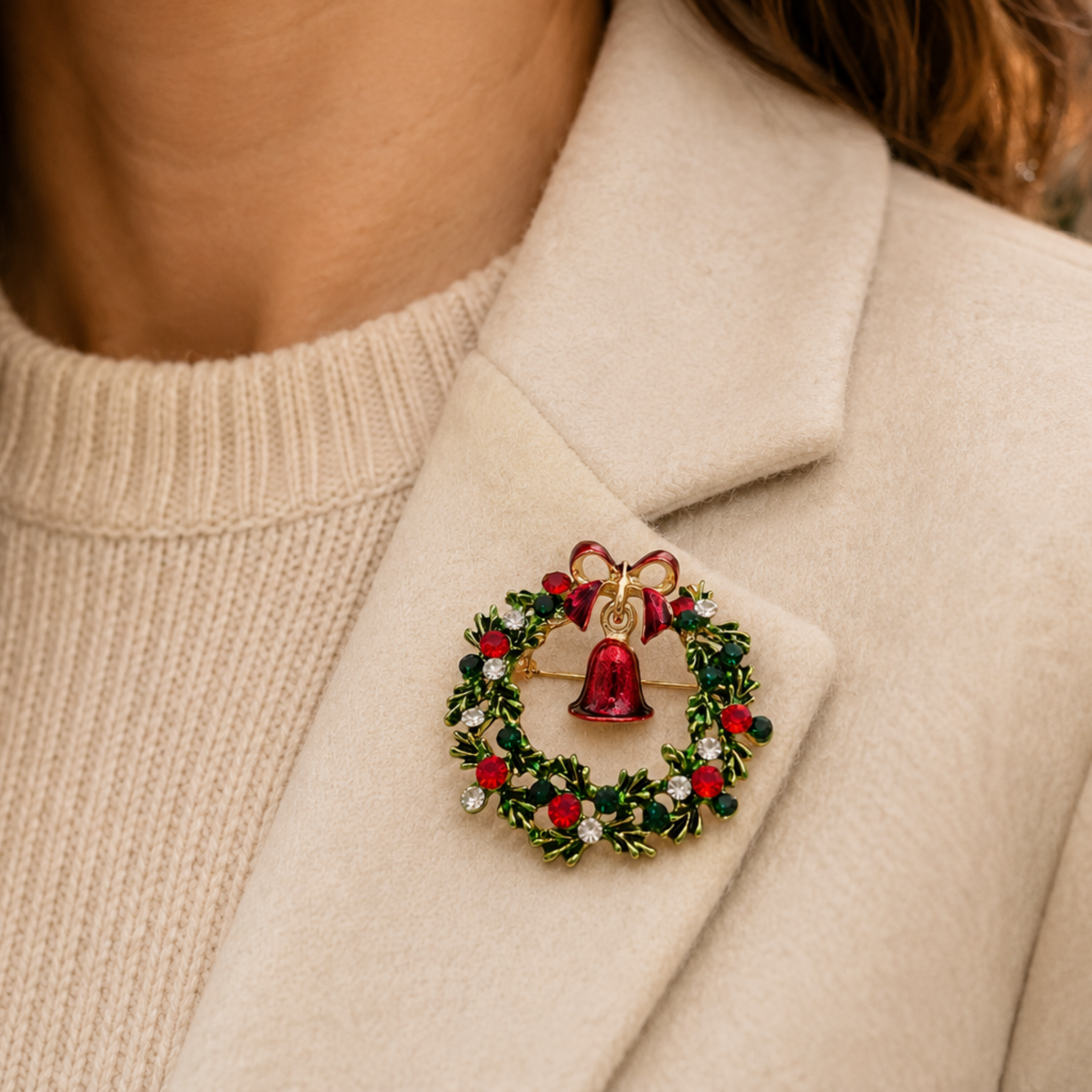 A close-up of a woman wearing a cream-colored coat and a ribbed cream sweater, featuring a festive Christmas wreath brooch pinned to the lapel. The brooch is detailed with green leaves, red berries, a red bow, and a gold bell.