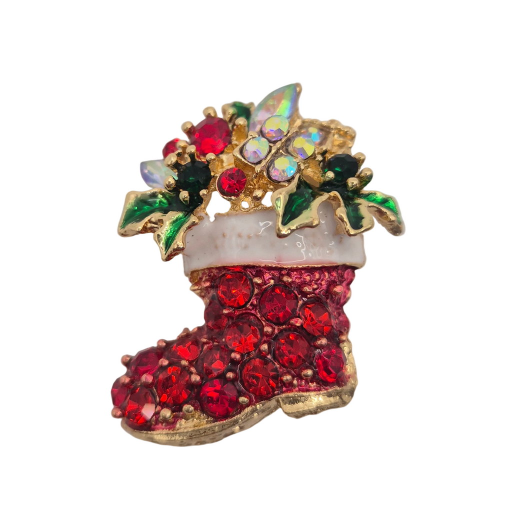 A Christmas stocking brooch featuring red rhinestones, gold-tone metal, green and white enamel leaves, and iridescent crystal accents, isolated on a white background.