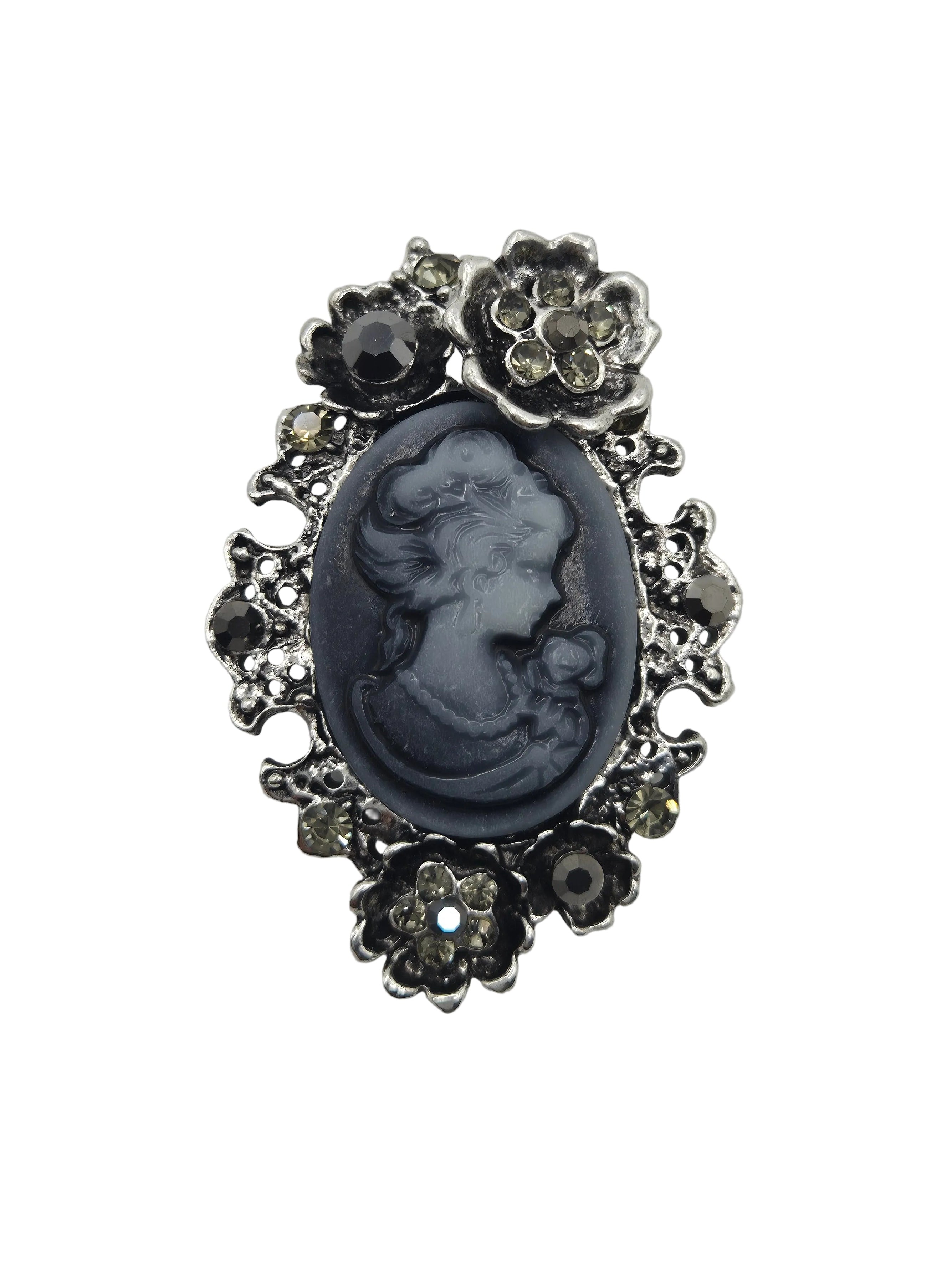 A vintage-style cameo brooch featuring a dark gray carved oval portrait in the center, surrounded by an ornate silver-tone floral border with small clear rhinestones, set against a plain white background.