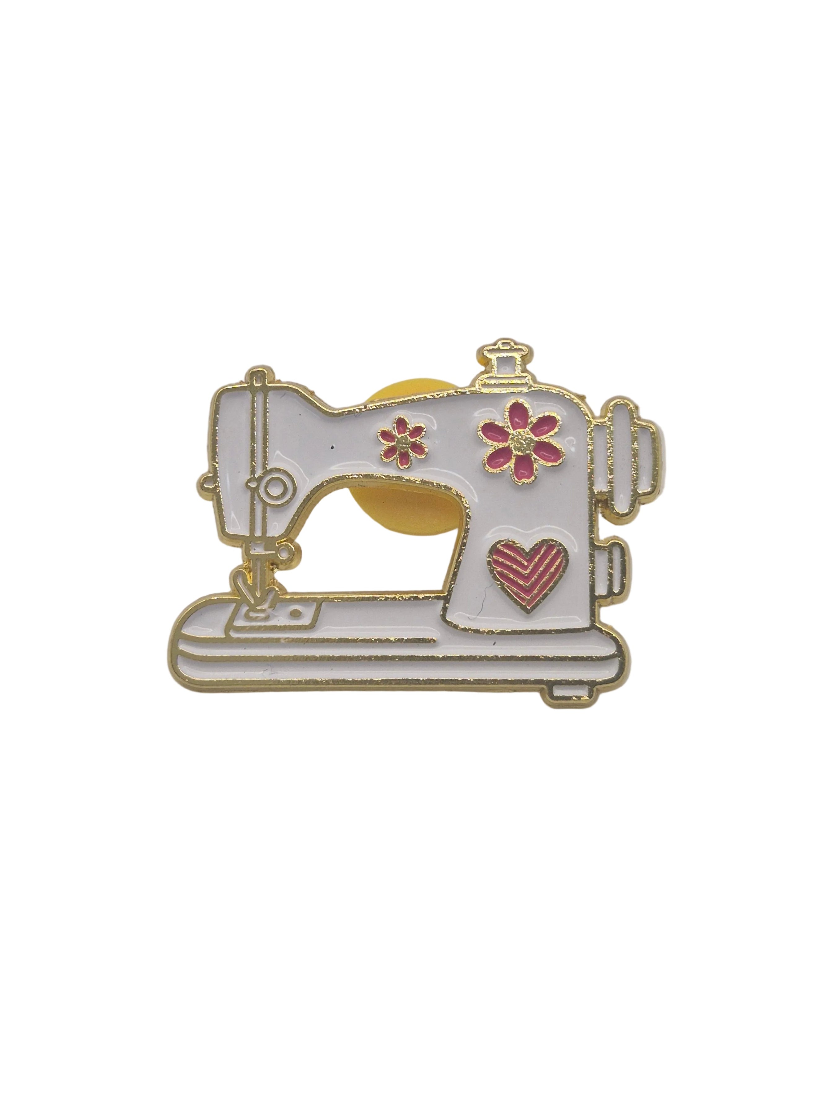 A small enamel pin shaped like a sewing machine, with a white body, gold-tone metal outlines, pink flower and heart details, and a yellow circular accent on a plain white background.