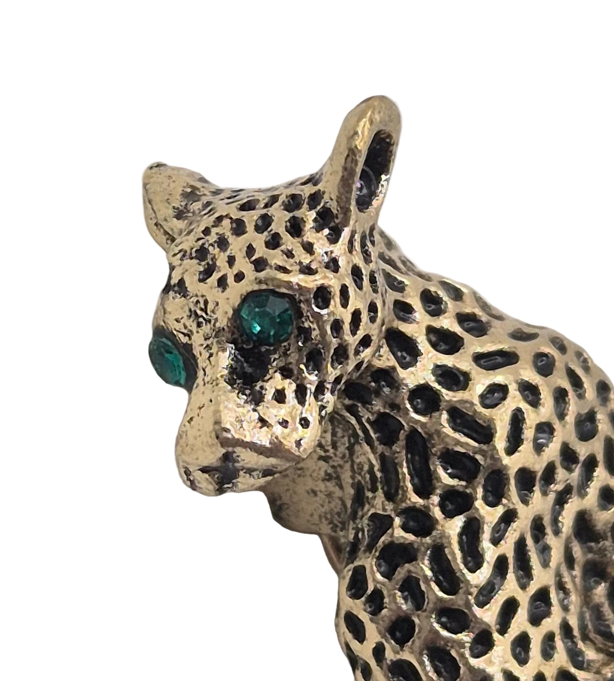 A close-up of a leopard brooch with a textured gold-toned body, black rosette spots, and green gemstone eyes, shown against a plain white background.
