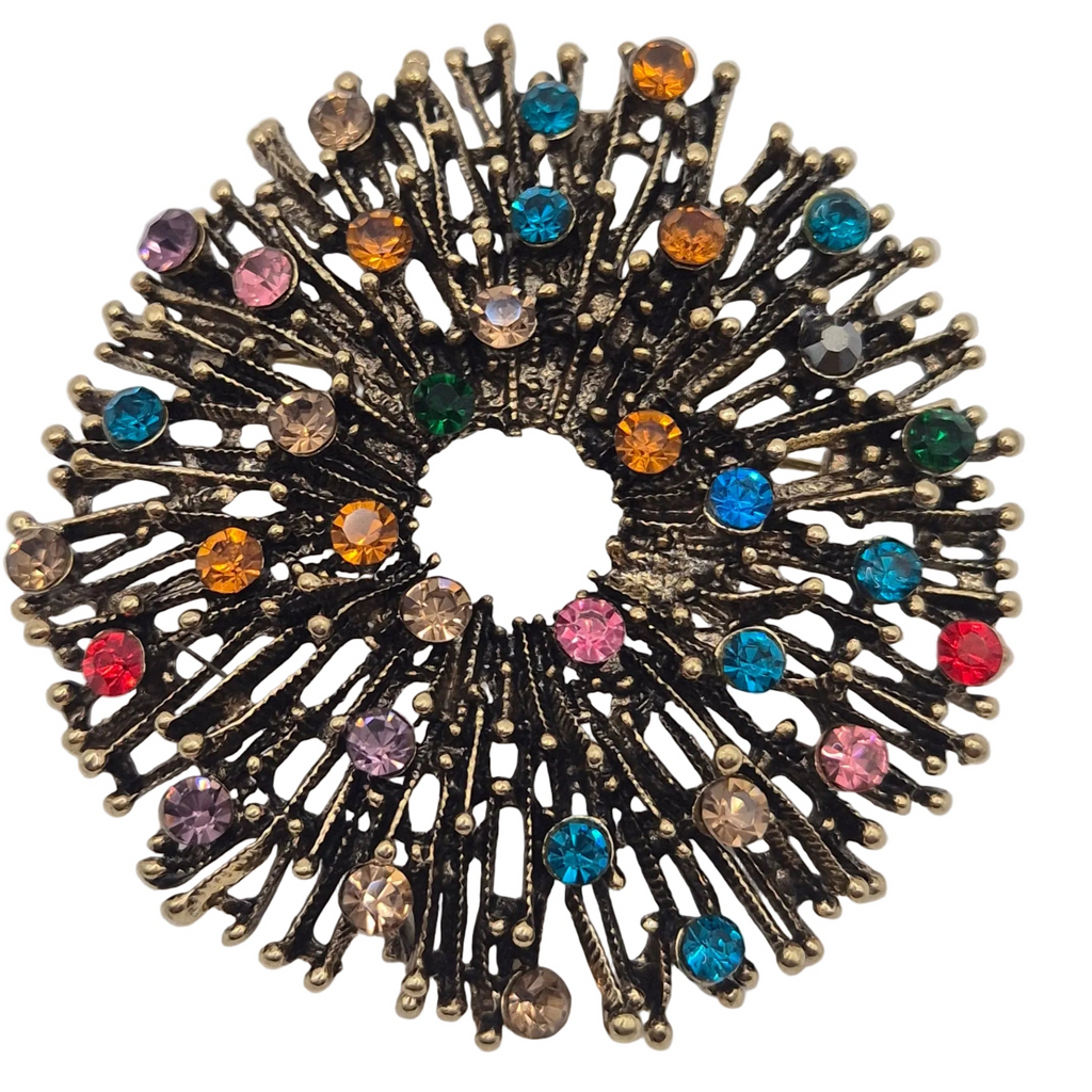A close up shot of a circular firework brooch featuring a antique gold metal base with multiple radiating spikes adorned with small, colourful rhinestones in pink, blue, orange, green, and purple, set against a plain white background.