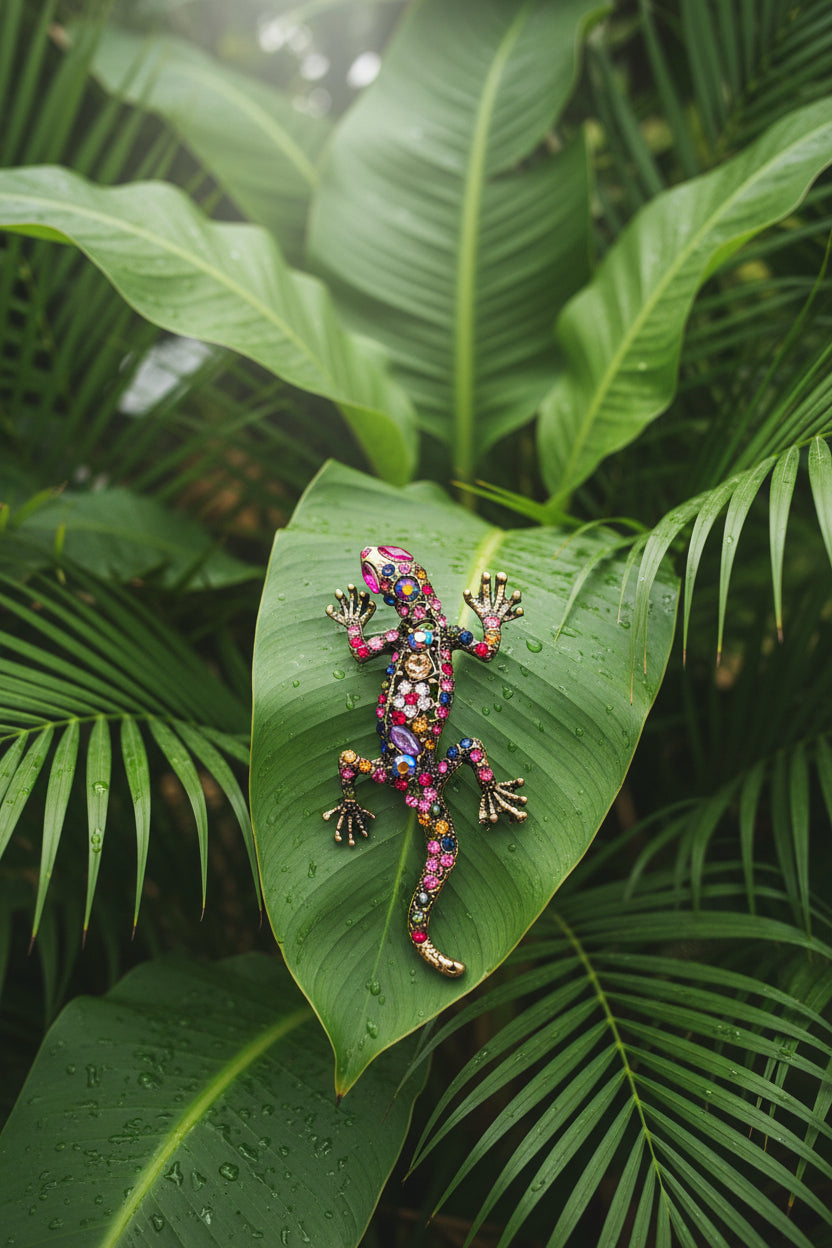 A decorative lizard or gecko-shaped brooch embellished with multicolored rhinestones and gold-tone metal, shown on a green leafy background.