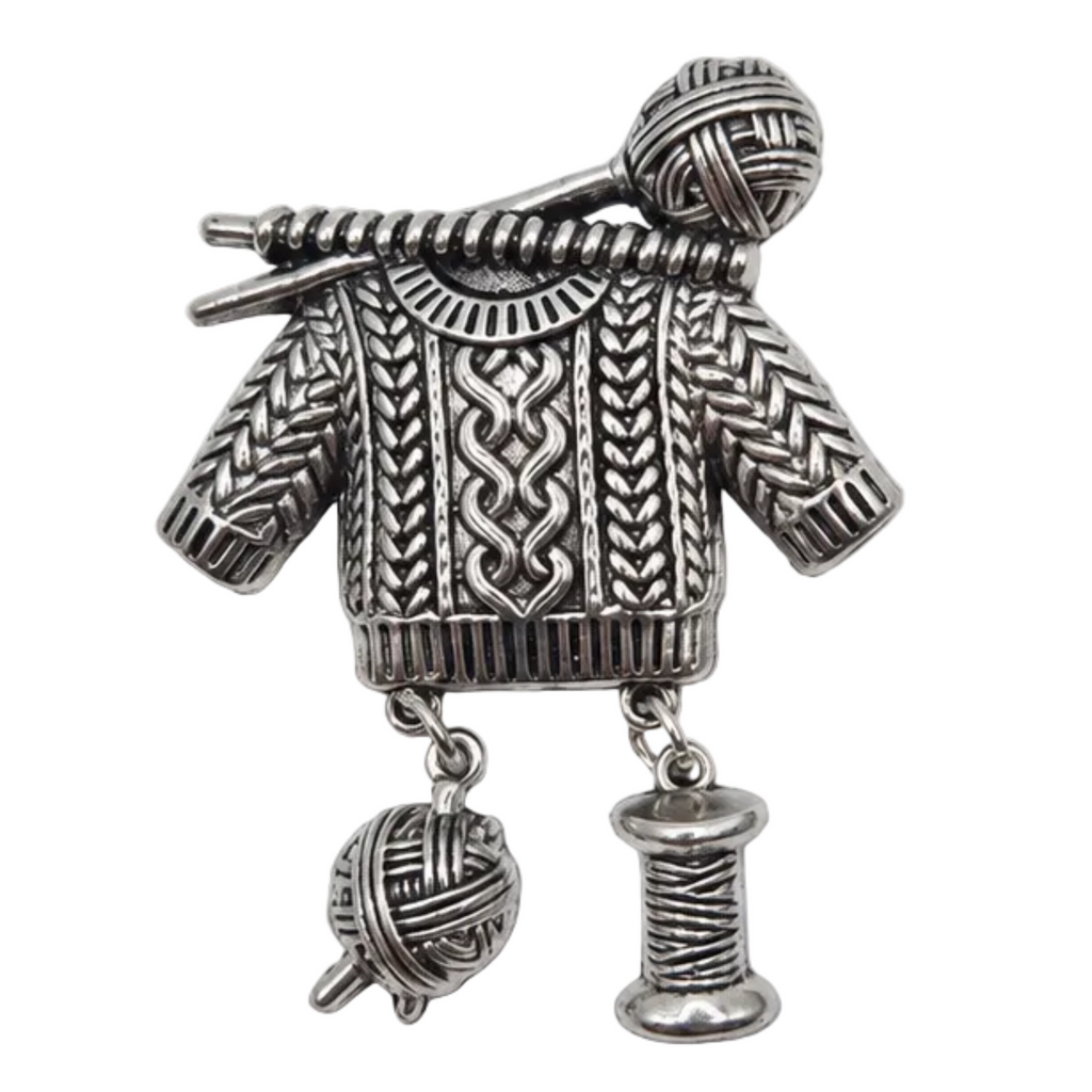 A silver-toned metal brooch shaped like a knitted sweater with knitting needles and yarn balls, shown on a plain white background.