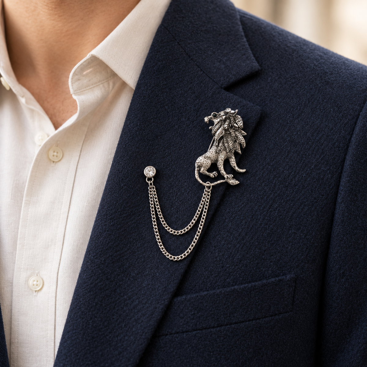 Silver-tone lion brooch with two draped chains and a round crystal pin, worn on a navy jacket lapel over a cream shirt.