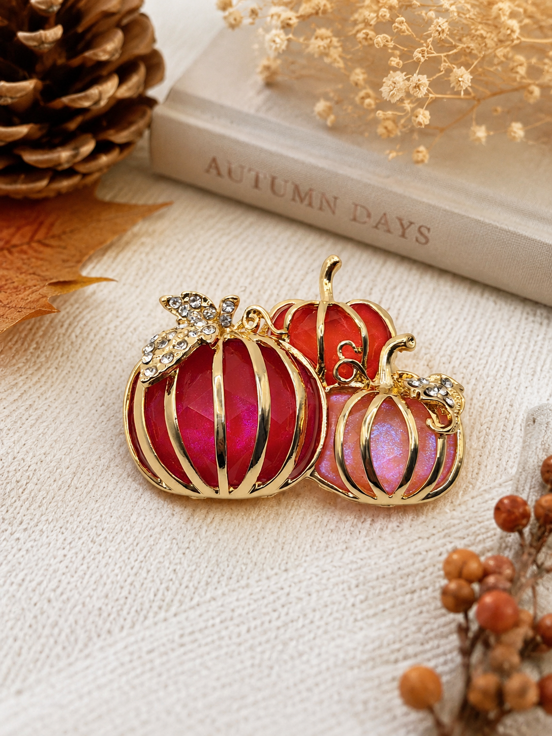 Two gold-plated pumpkin-shaped brooches with red and pink iridescent enamel, decorated with small clear rhinestones on the stems, displayed on a textured cream fabric with autumn-themed props.