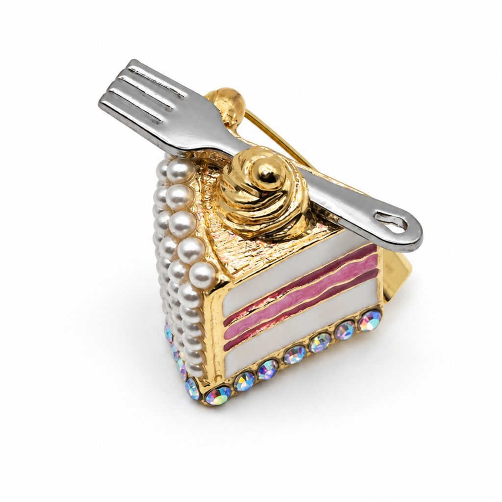 A brooch shaped like a slice of cake with a fork on top, featuring gold-tone metal, white faux pearls, pink enamel stripes, and iridescent rhinestones on a white background.