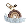 A bohemian-style woven rainbow keychain with a gold-tone lobster clasp, a small black enamel flower charm, and white tassels at the bottom, shown on a plain white background.