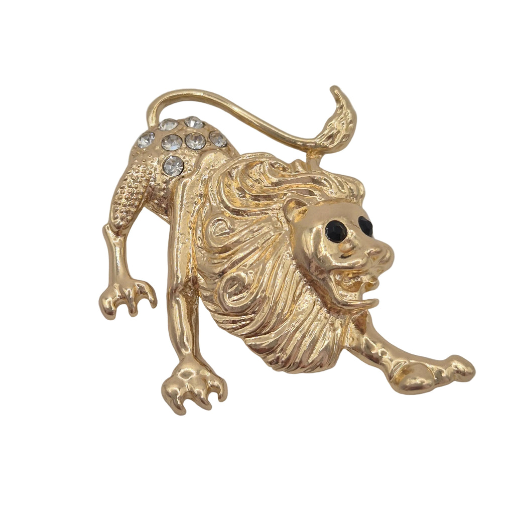 A gold lion figurine brooch with a textured mane, black eyes, embellished with clear rhinestones on its hindquarters, shown on a plain light gray background.