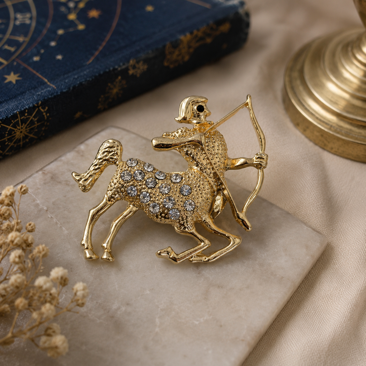 A gold-tone Sagittarius zodiac sign brooch decorated with clear rhinestones, displayed on a beige stone slab with a navy blue book and dried flowers in the background.