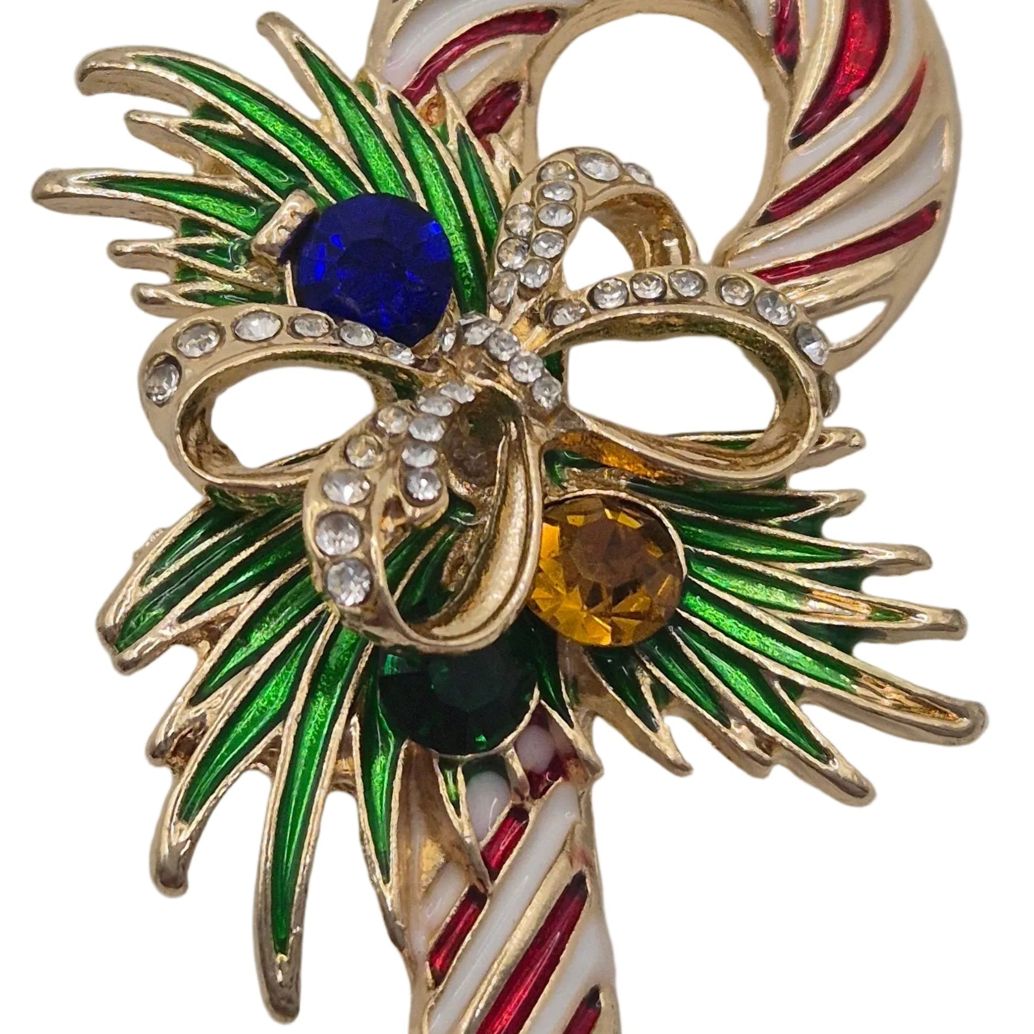 A close-up of a gold-tone Christmas ornament or brooch shaped like a candy cane with a bow, featuring green enamel leaves, red and white striped details, and faceted gemstones in blue, amber yellow, and dark green.