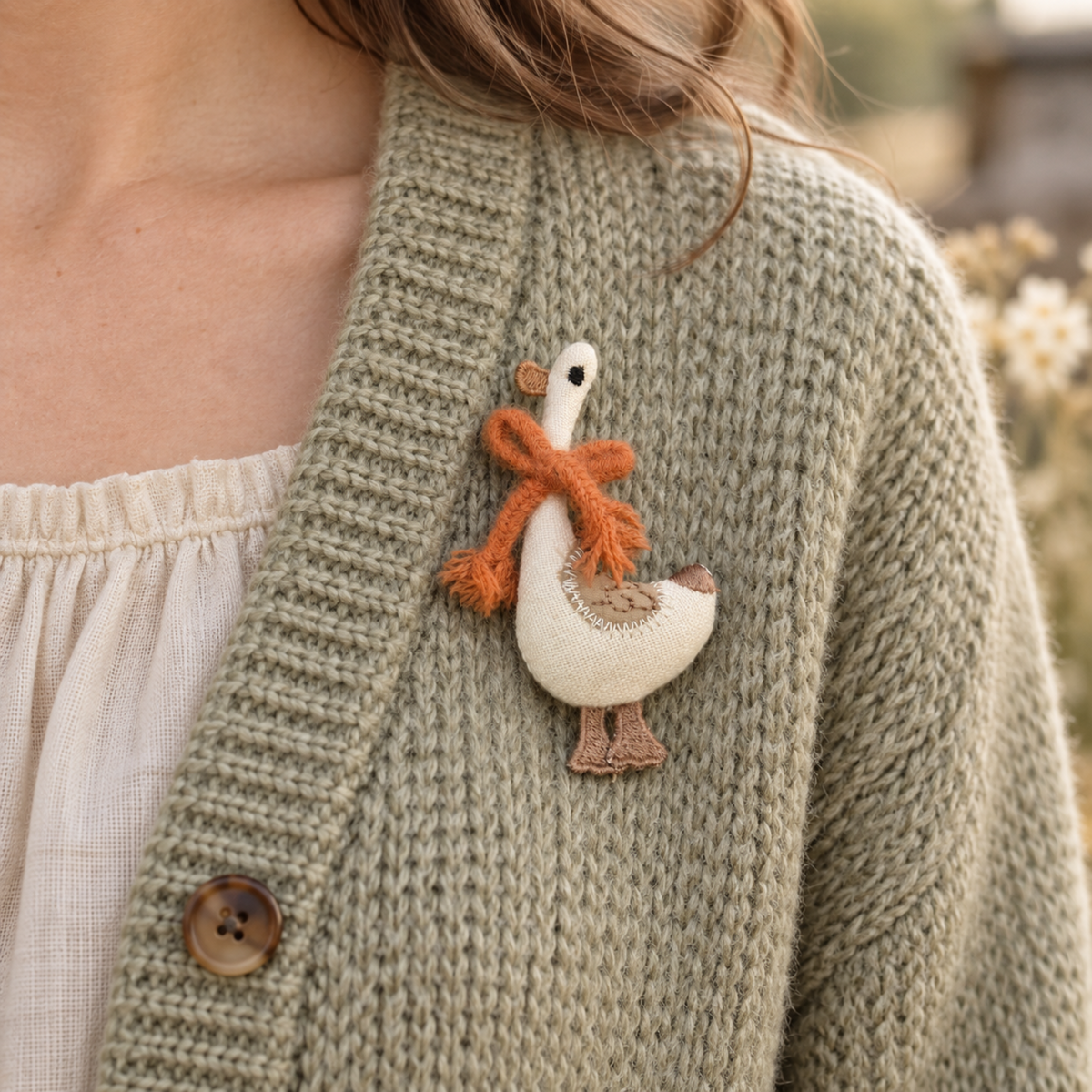 A close-up of a person wearing a chunky knit cardigan in a muted sage green colour, featuring a cream-coloured fabric goose pin with an orange scarf and brown details on the chest.