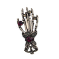 A gothic-style skeleton hand brooch featuring silver-toned metal with dark oxidized details, accented by red gemstones and small clear rhinestones, shown on a plain white background.