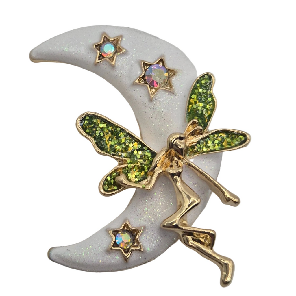 A whimsical fairy and crescent moon brooch featuring a gold-tone fairy with glittery green wings flying over a white glittery crescent moon, accented with three iridescent star rhinestones.