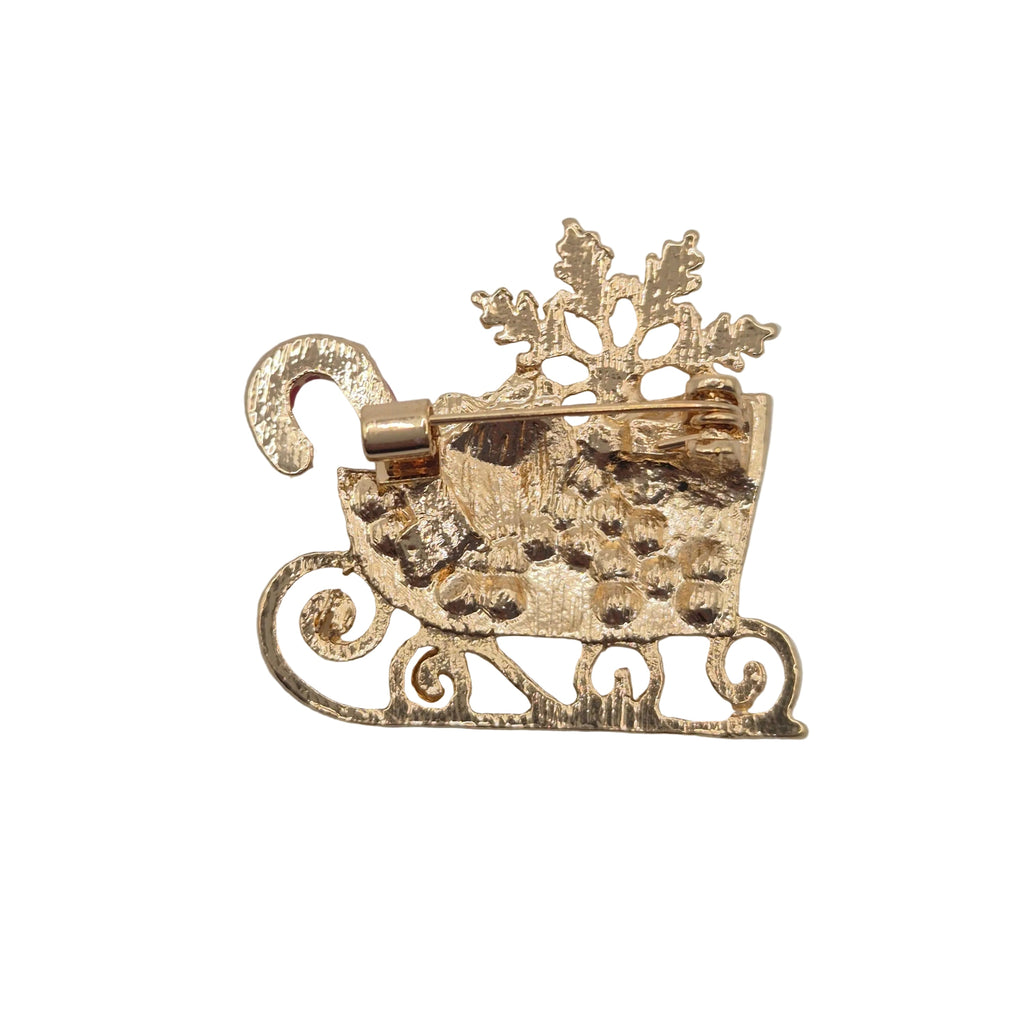 A gold-toned Christmas-themed brooch shaped like a sleigh carrying a candy cane and snowflake, photographed on a white gray background.