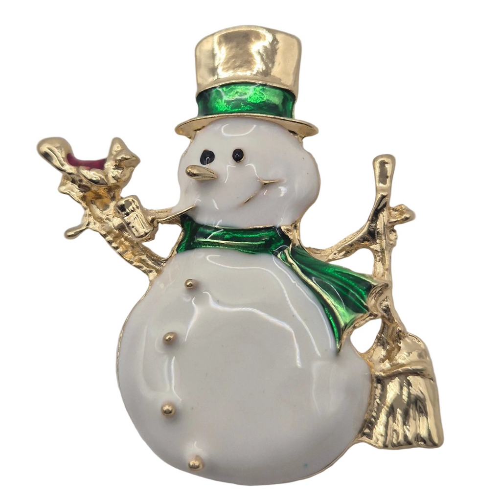 A snowman-shaped brooch or pin with a glossy white enamel body, gold-tone metal trim, a green hat band and scarf, and small red accent details on the hat and arm.
