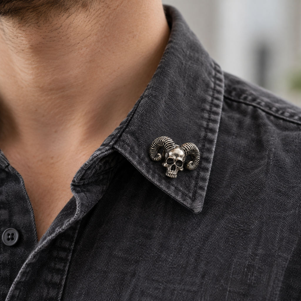 Small silver-tone skull brooch with curled ram horns pinned to the collar of a charcoal shirt, showing a casual Gothic styling idea.