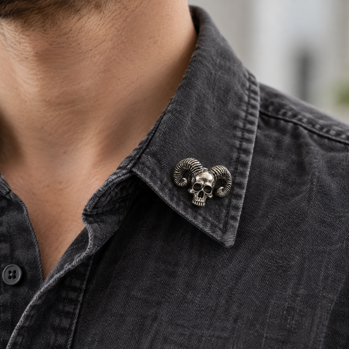 Small silver-tone skull brooch with curled ram horns pinned to the collar of a charcoal shirt, showing a casual Gothic styling idea.