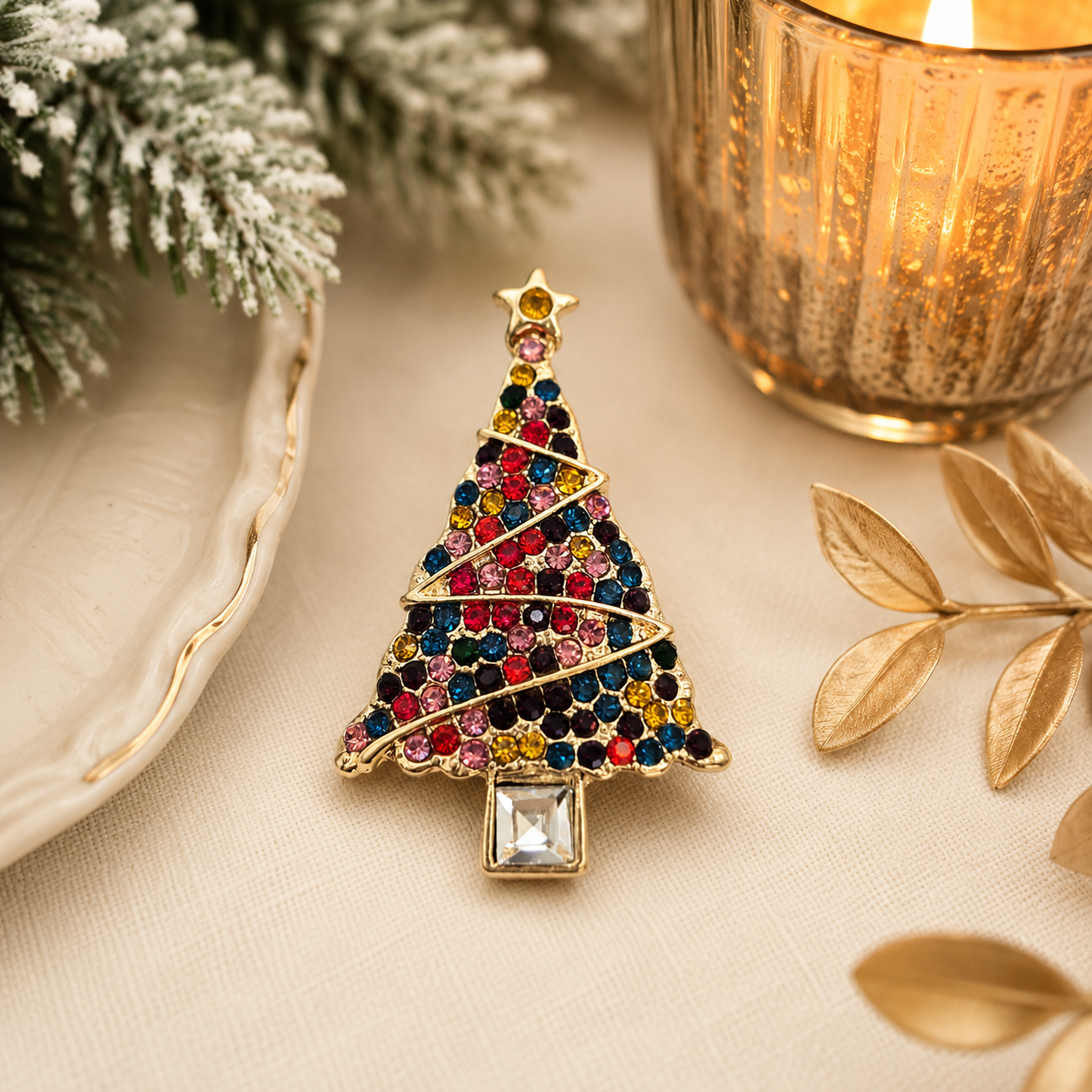 Colourful crystal Christmas tree brooch with a gold-tone finish displayed on soft cream fabric, surrounded by frosted greenery, a glowing candle and elegant festive décor, creating a warm and sophisticated holiday lifestyle image.