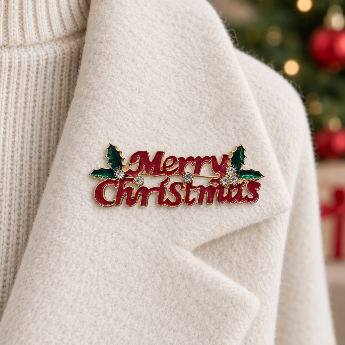 A close-up of a red enamel lapel pin reading 'Merry Christmas' with green holly leaves and small rhinestone accents, pinned onto a cream-colored knit sweater. A blurred Christmas tree and gift box are visible in the background.