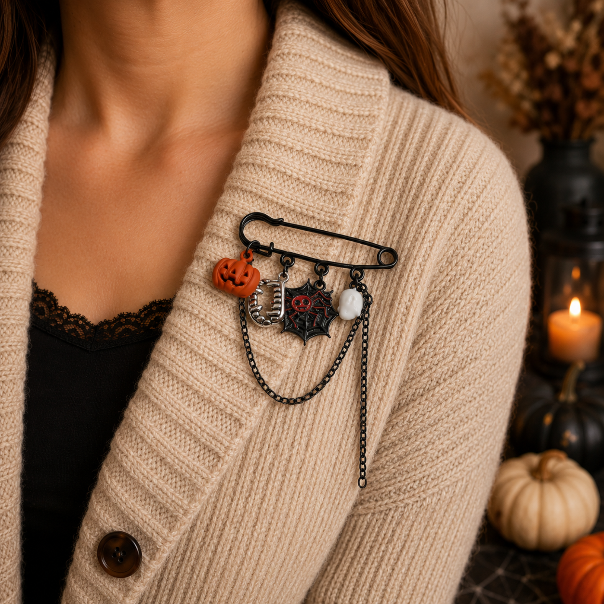 A close-up of a person wearing a black safety pin brooch decorated with Halloween-themed charms, including a small orange pumpkin, a spider, and a white skull, pinned to a beige knit cardigan.