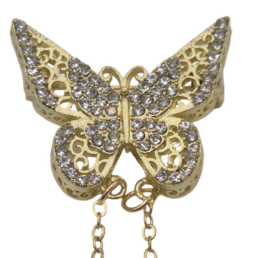 Gold butterfly-shaped brooch or pin embellished with clear rhinestones, featuring an openwork filigree design and a dangling chain, shown on a plain white background.