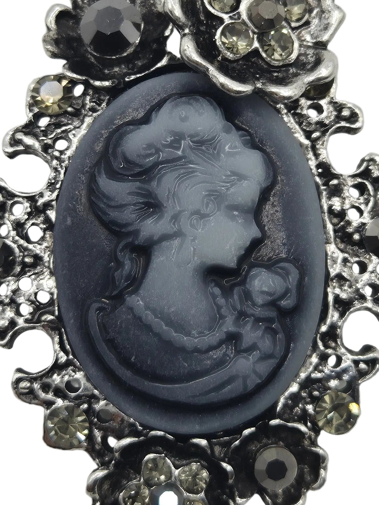 A close-up of a vintage-style cameo brooch featuring a dark gray carved oval profile of a woman with a rose, surrounded by an ornate silver-tone filigree border with clear rhinestones.