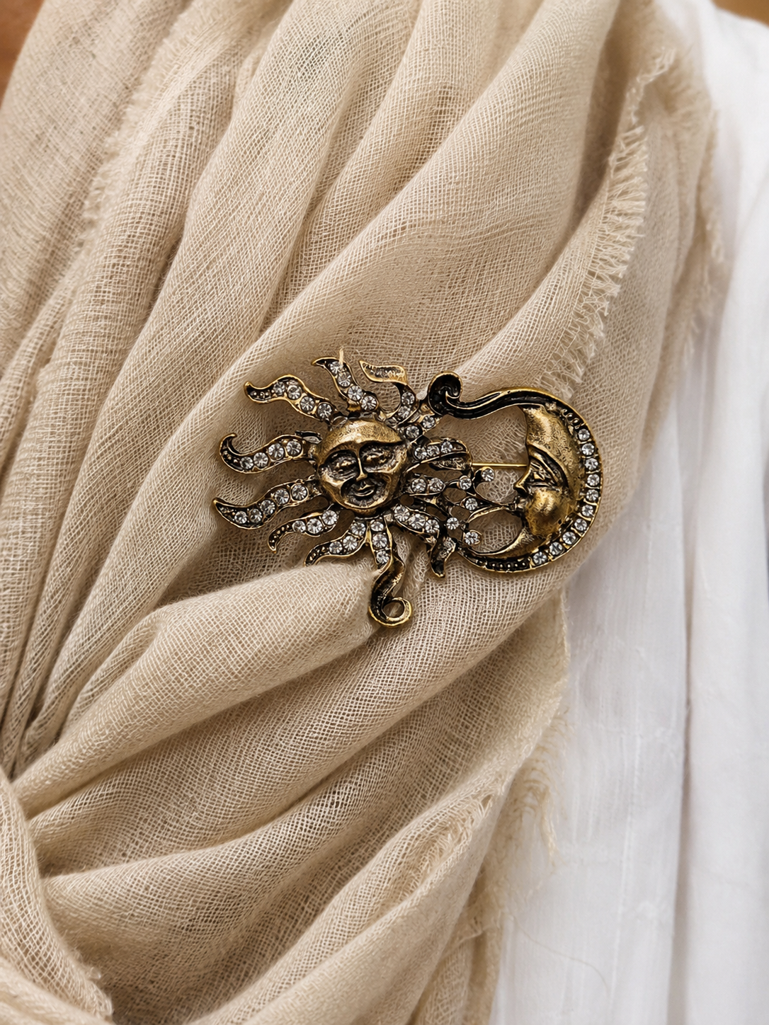 An antique gold-toned brooch featuring a sun and crescent moon design, embellished with small clear rhinestones, pinned onto a beige textured scarf.
