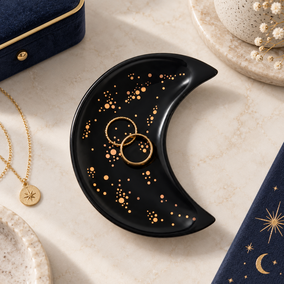 Black ceramic crescent moon trinket dish with gold celestial detailing, styled with two rings on a cream dressing table with navy and gold accessories.