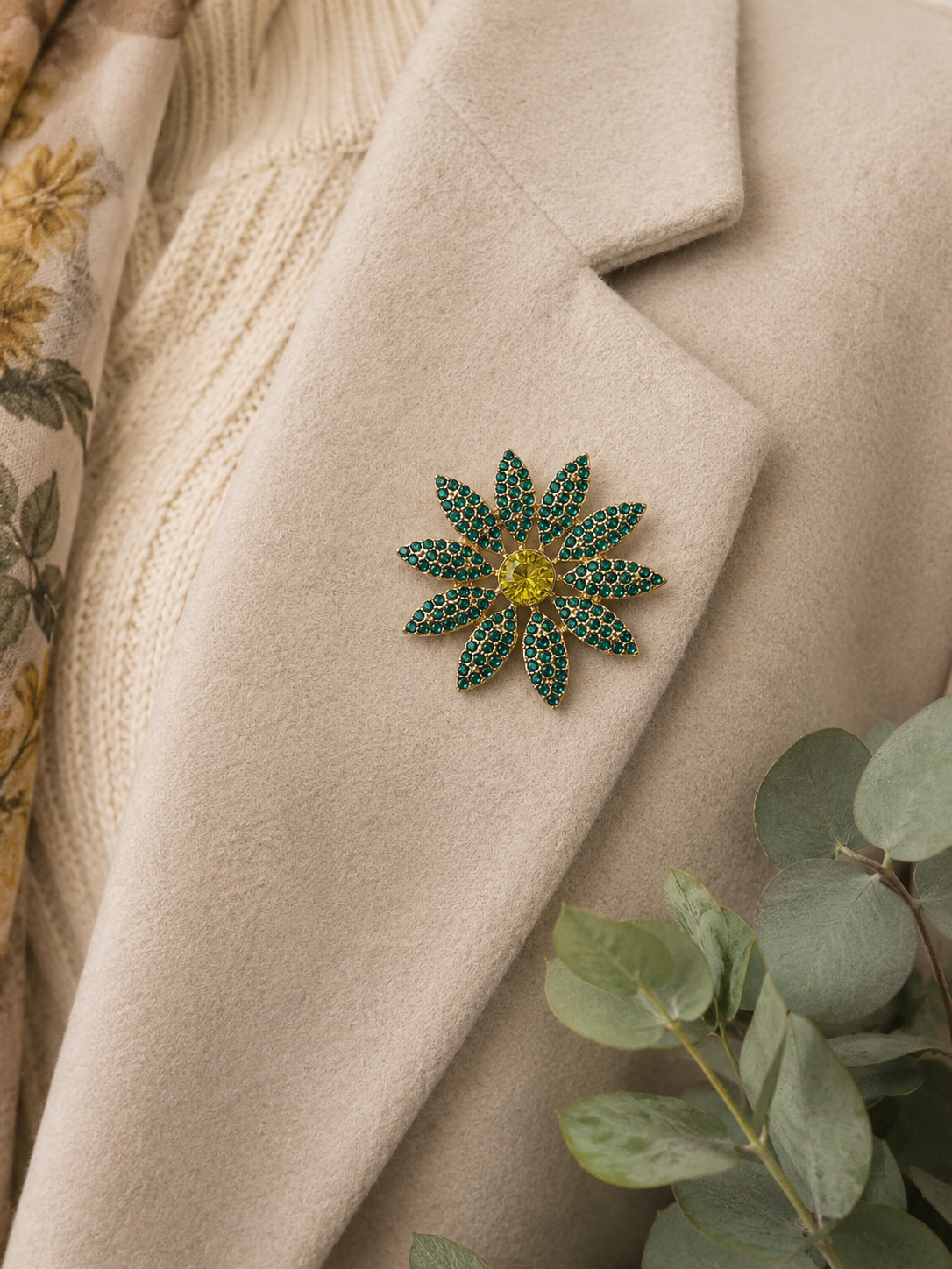 A flower-shaped brooch with green and gold rhinestones pinned onto a beige wool coat, styled with a cream knit sweater and eucalyptus leaves.