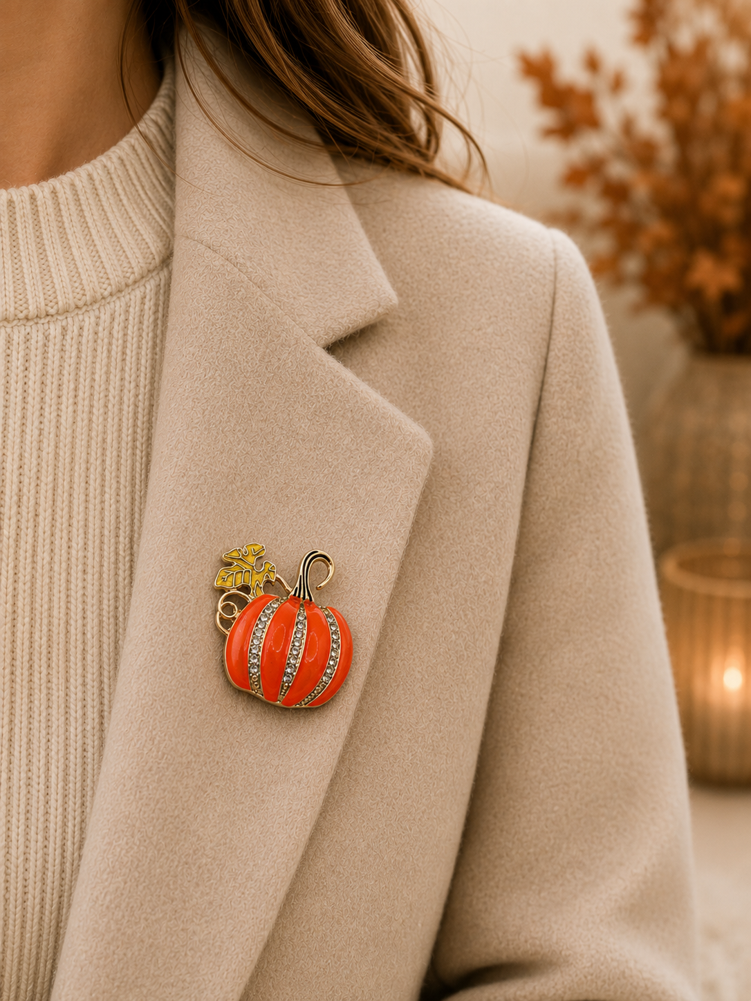 A close-up of a person wearing a beige coat over a cream ribbed sweater, with a small pumpkin-shaped brooch pinned to the left lapel. The brooch is orange with vertical white stripes and a gold stem and leaf detail.