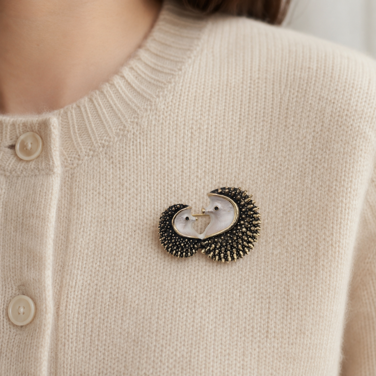 A close-up of a person wearing a cream-coloured knit cardigan with a double hedgehog brooch pinned to the chest.