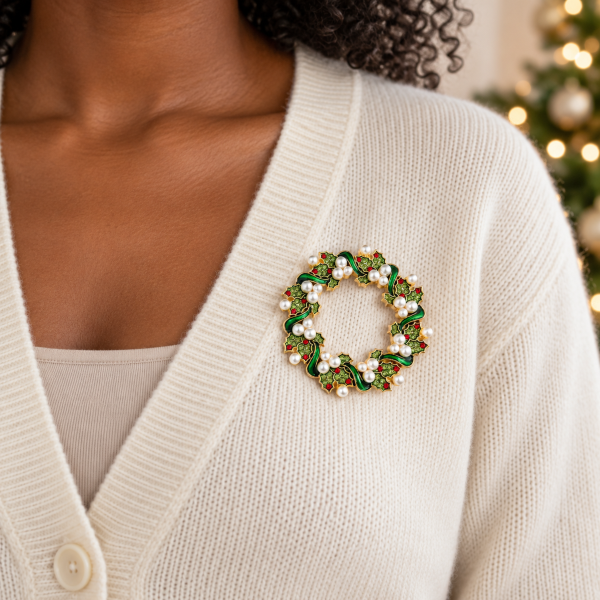 A close-up of a woman wearing a holiday wreath brooch on her cream-colored cardigan sweater. The brooch features green enamel leaves, gold-tone metal outlines, and small white faux pearls.