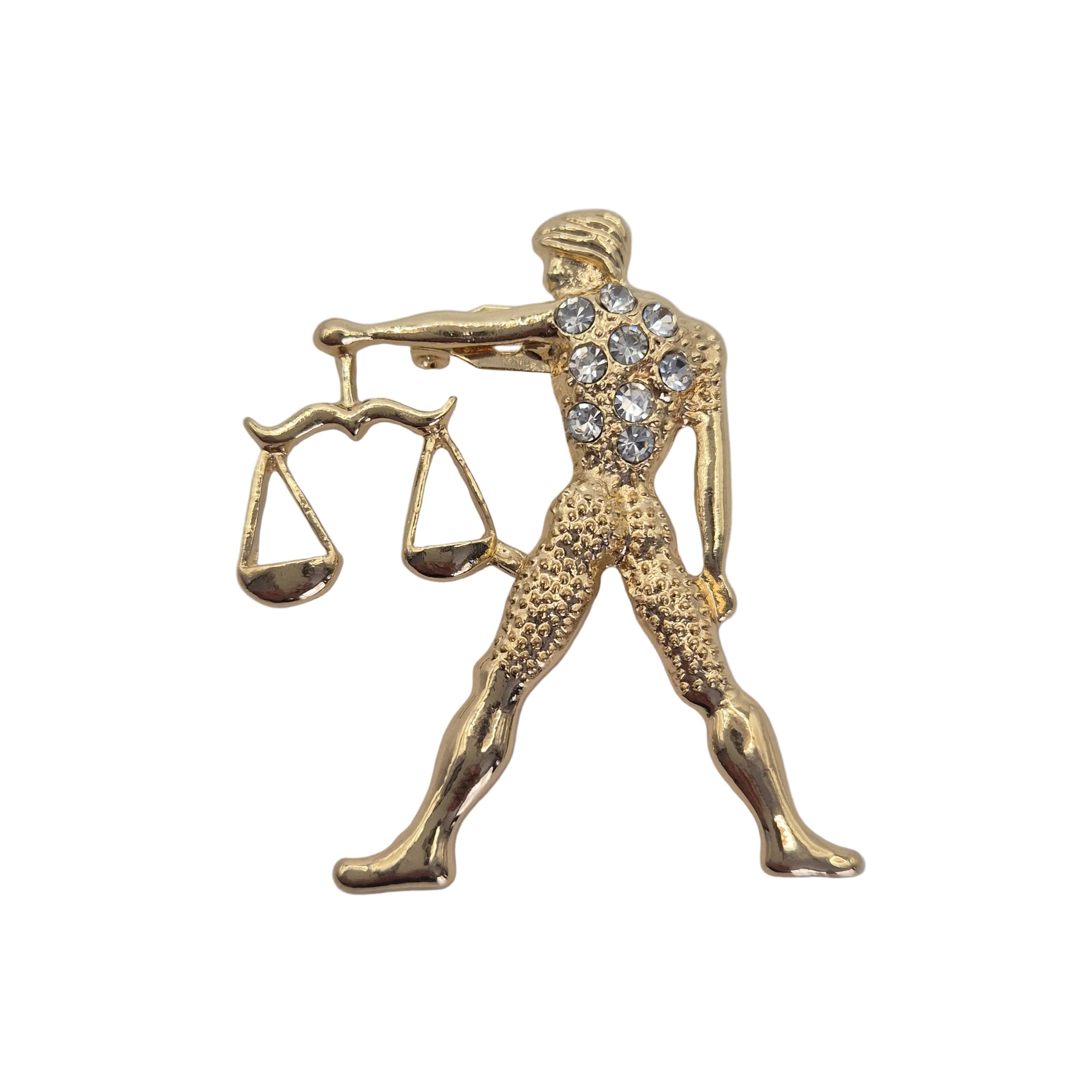 A gold-tone Libra zodiac sign pin featuring a blindfolded figure holding a balance scale, with clear rhinestones on the chest, set against a plain white background.