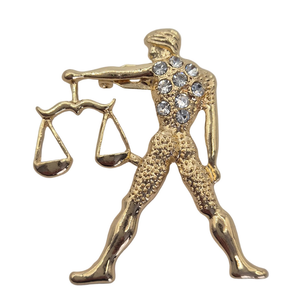 A gold-tone metal pin shaped like the zodiac sign of Libra, featuring a figure holding a balance scale and adorned with clear rhinestones on the chest, set against a plain white background.