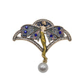 An Art Deco style brooch featuring a fairy design with blue enamel wings, blue rhinestone accents, a gold vine stem, and a dangling white pearl, shown on a plain white background.
