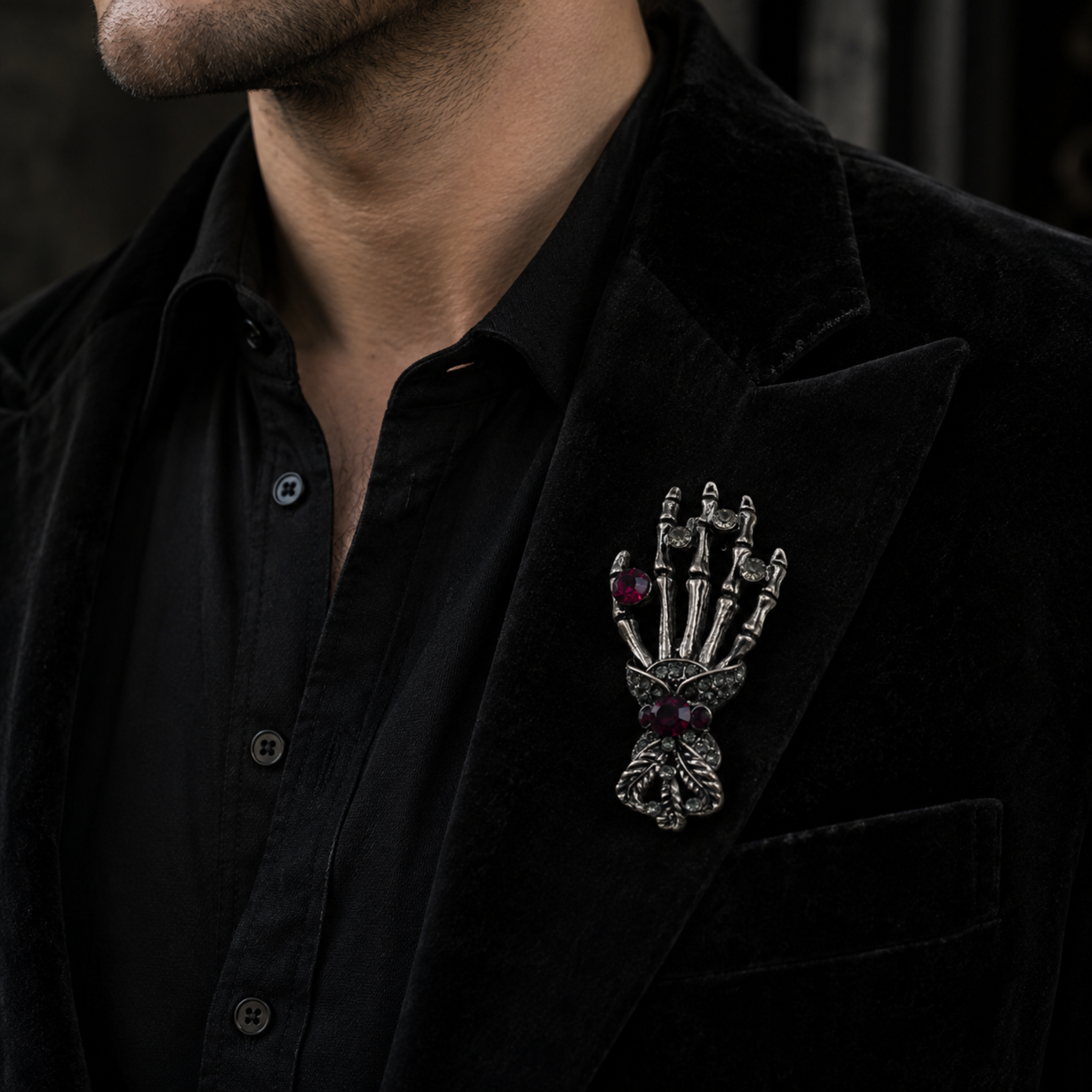 A gothic-style skeleton hand brooch with dark red gemstone eyes and fingers, pinned to the lapel of a man wearing a black velvet blazer and black button-down shirt.