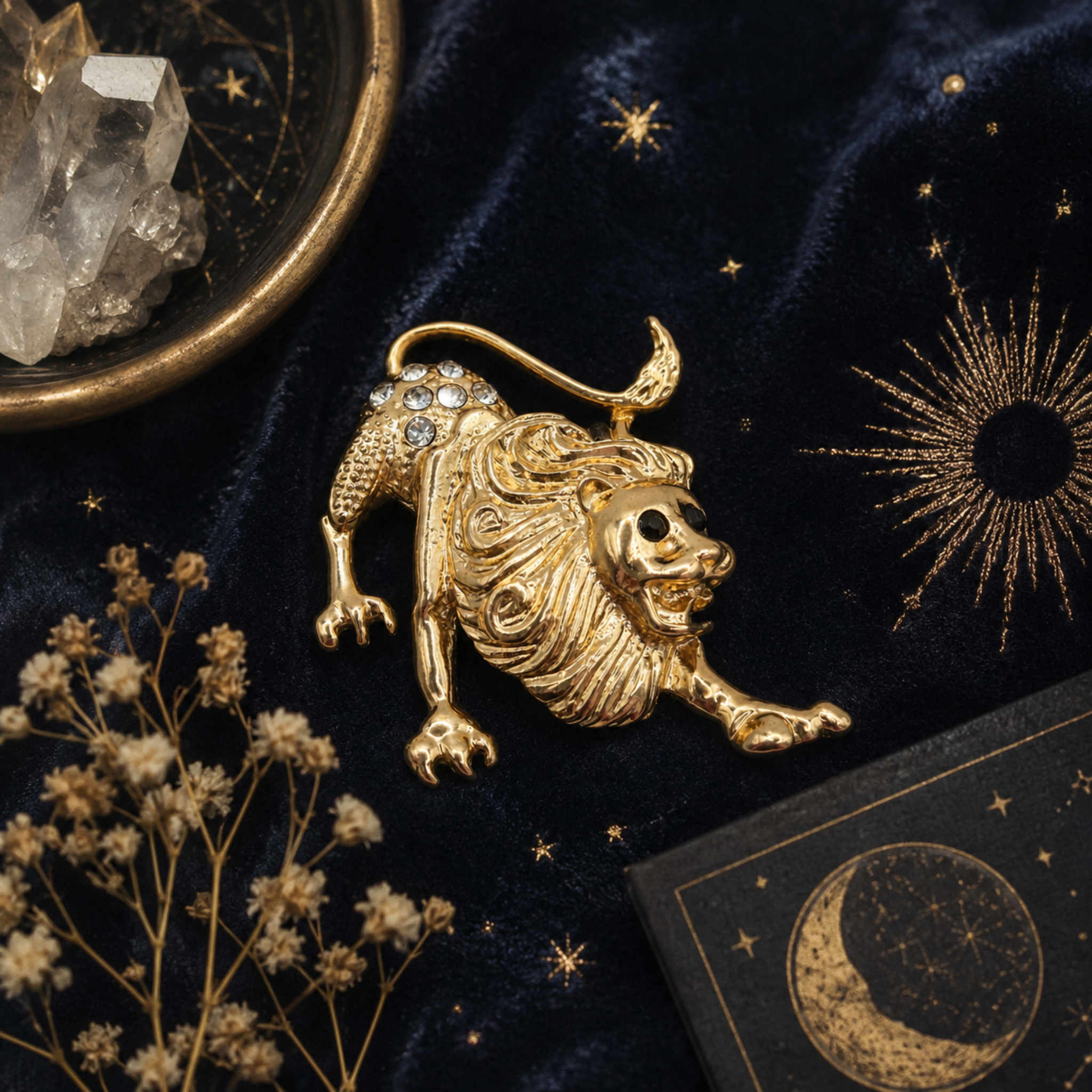 A gold lion-shaped brooch with a skull face and rhinestone-spined tail, displayed on a dark velvet fabric with gold celestial embroidery, dried flowers, a crystal cluster, and a card featuring a moon and stars.