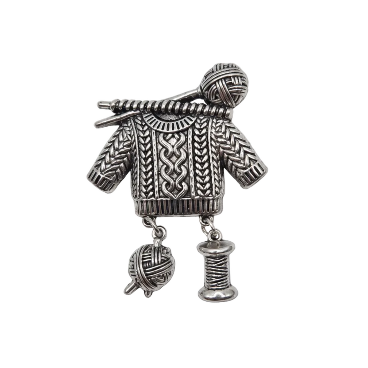 A silver-toned metal brooch shaped like a knitted sweater with knitting needles, yarn balls, and a spool of thread, displayed on a plain white background.