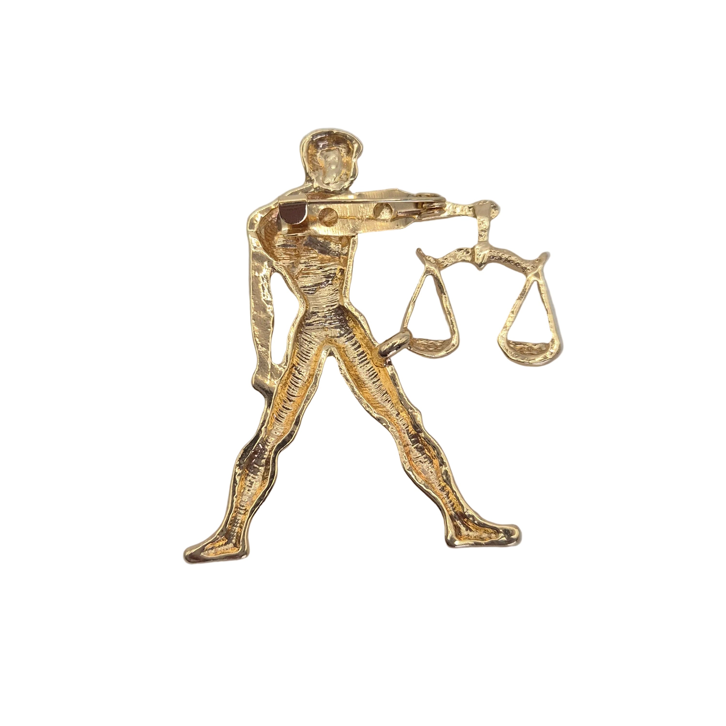A gold-tone metal pin shaped like the zodiac sign Libra, depicting a figure holding a balance scale, on a plain light gray background.