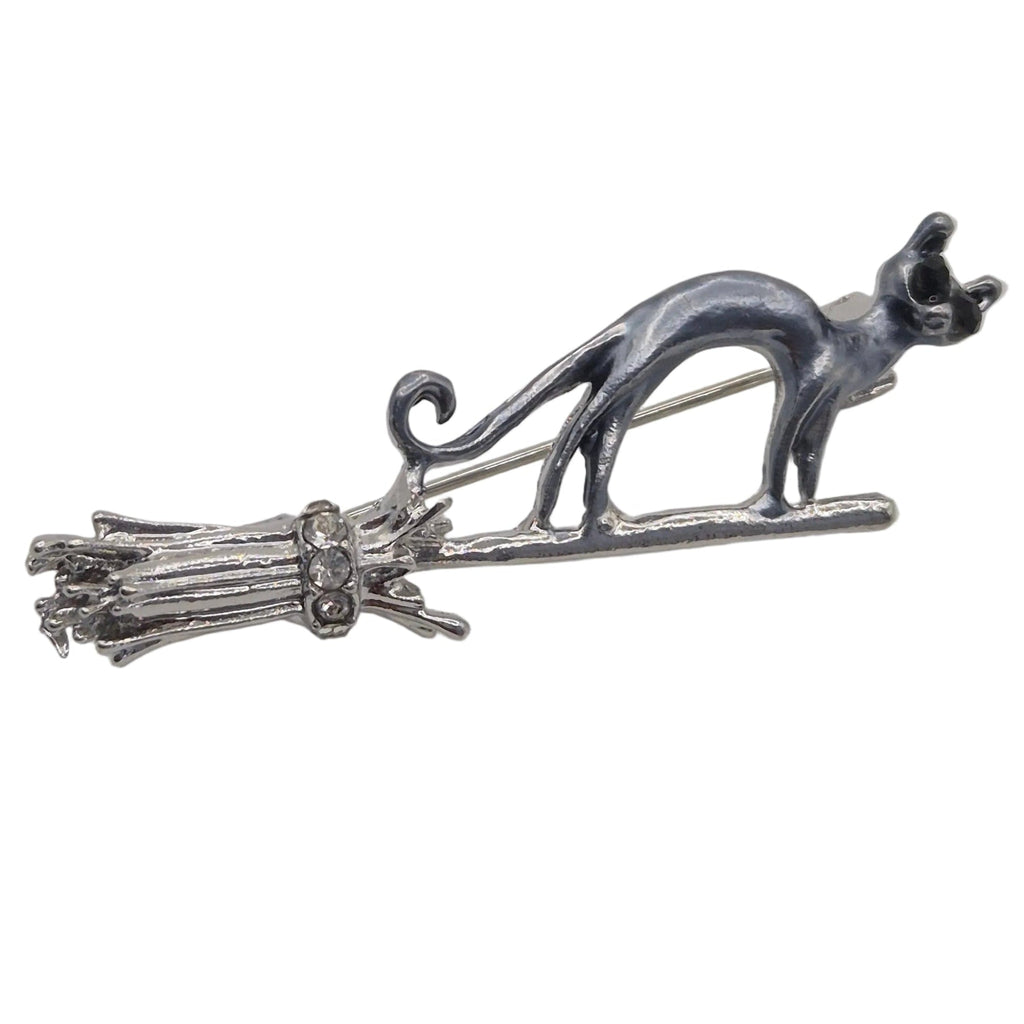 A silver and grey toned metal brooch featuring a cat standing on a broomstick, photographed against a plain white background.