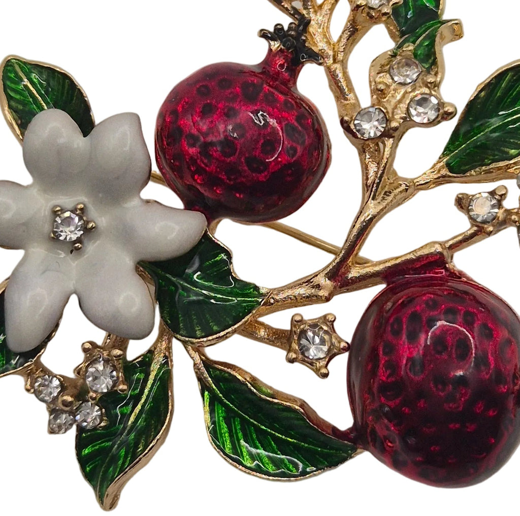 A gold-tone brooch featuring two glossy dark red pomegranate fruits, a white enamel flower, and green enamel leaves, accented with small clear rhinestones on a plain white background.