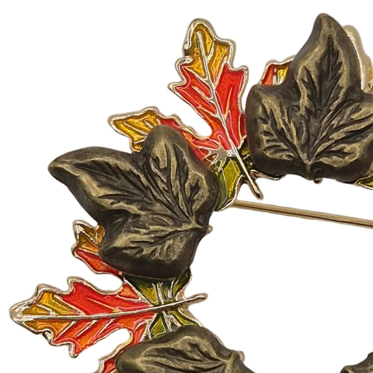 A close-up of a vintage-style metal wreath or brooch featuring textured dark bronze leaves with red, orange, and yellow enamel accents on a white background.