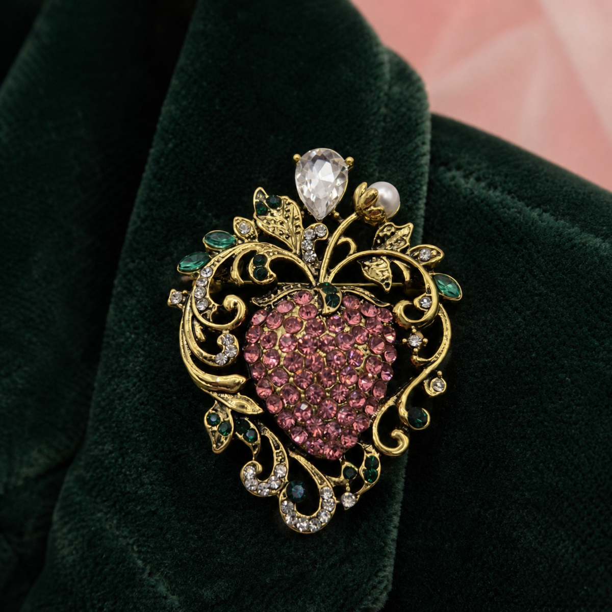An ornate gold brooch featuring a heart-shaped center paved with pink stones, surrounded by green and clear rhinestones, a pearl, and a clear teardrop gem, displayed on a dark green velvet surface.