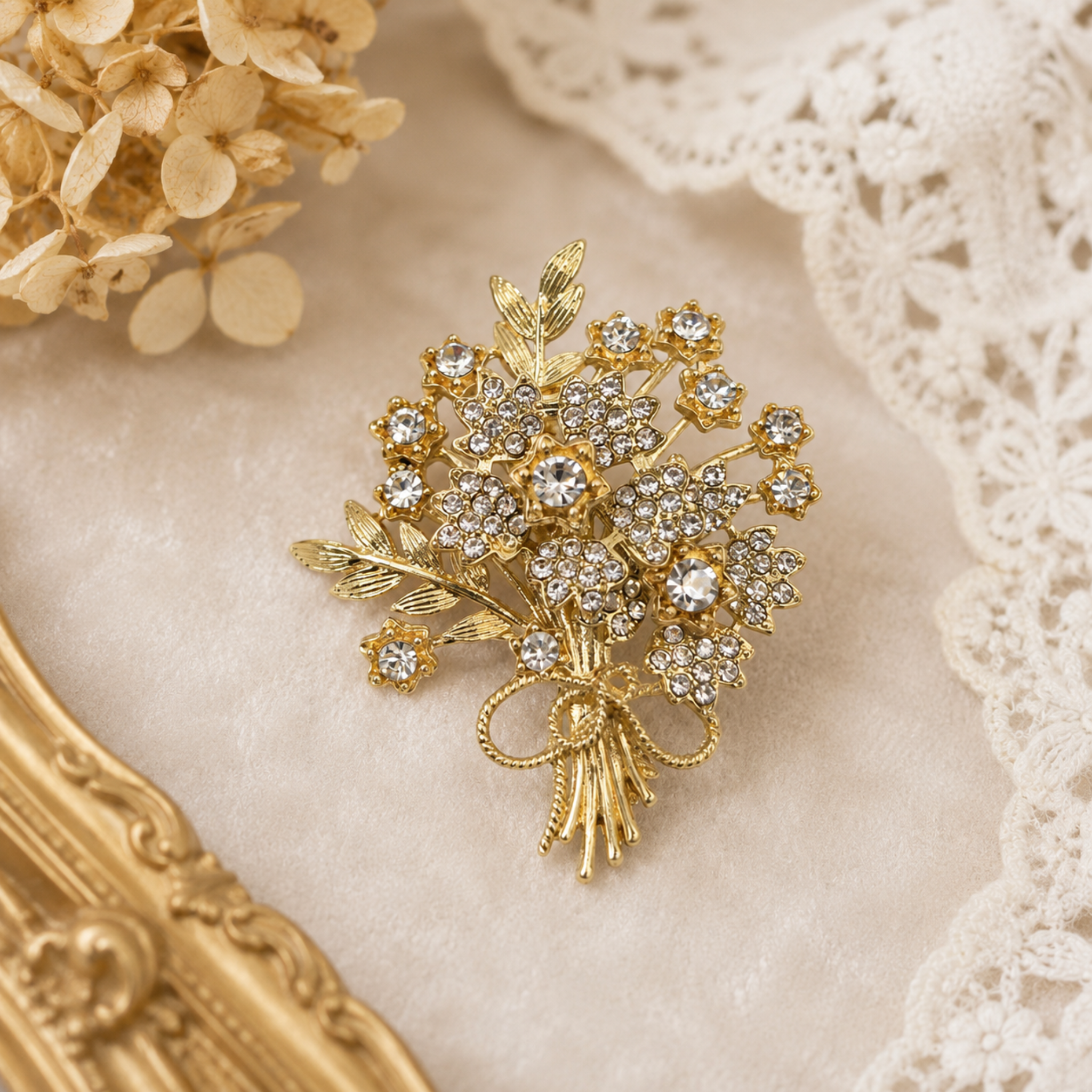 A gold-toned brooch shaped like a bouquet of flowers, adorned with clear rhinestones, displayed on a soft cream fabric with lace and dried flowers in the background.