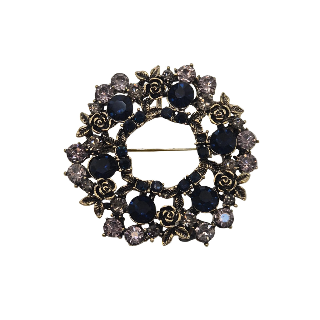 A vintage-style brooch featuring a circular design with dark blue faceted gemstones, antiqued gold-toned rose motifs, and clear rhinestones, photographed on a plain light gray background.