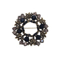 A vintage-style brooch featuring a circular design with dark blue faceted gemstones, antiqued gold-toned rose motifs, and clear rhinestones, photographed on a plain light gray background.
