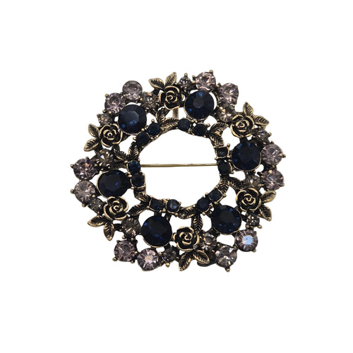 A vintage-style brooch featuring a circular design with dark blue faceted gemstones, antiqued gold-toned rose motifs, and clear rhinestones, photographed on a plain light gray background.