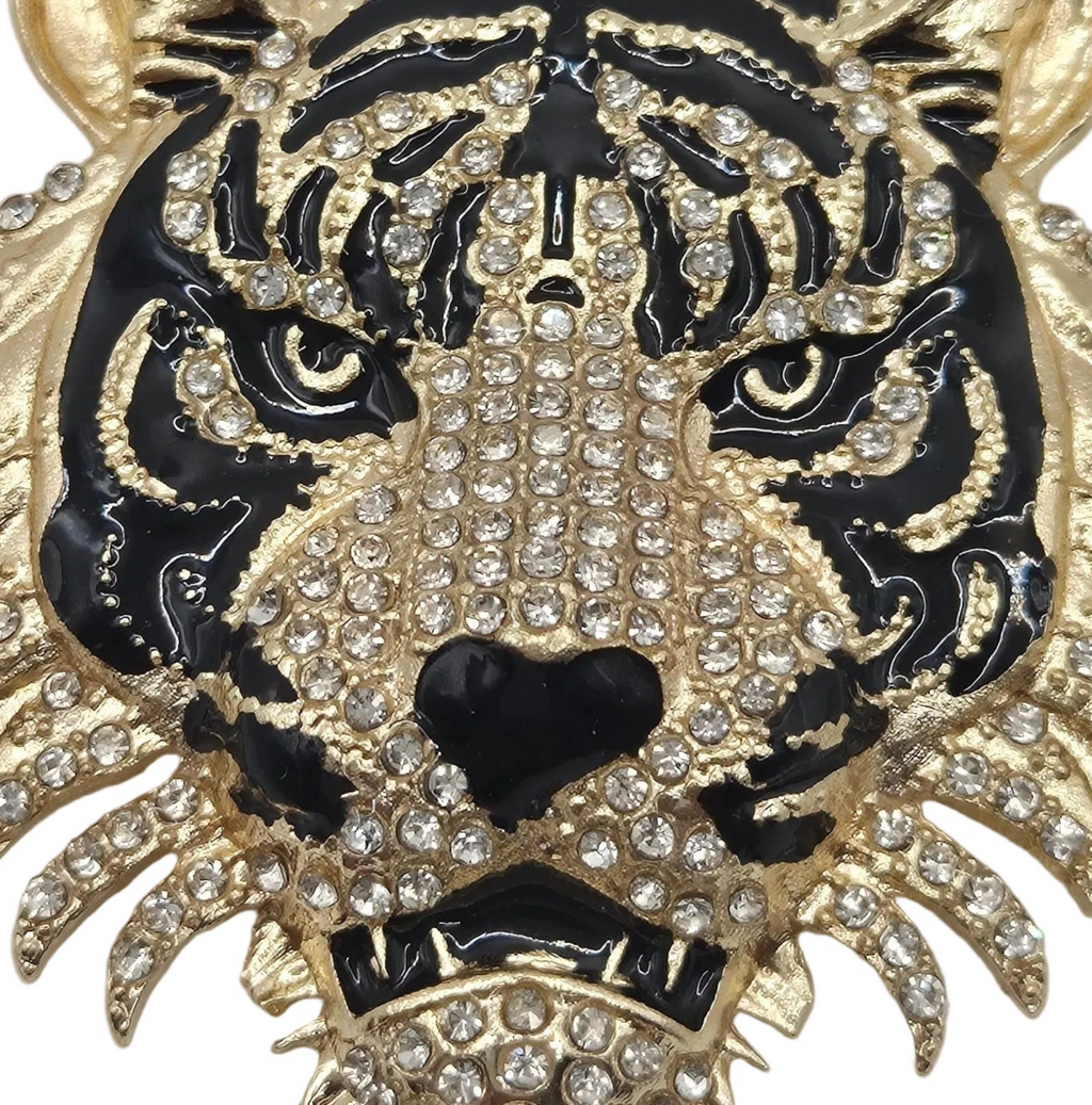A close-up of a tiger face brooch or pin featuring black enamel, gold-tone metal outlines, and rows of clear rhinestones across the forehead, eyes, nose, and surrounding details.