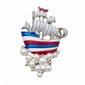 A silver-toned ship brooch featuring white sails, red and blue striped hull details, and a cluster of white pearls at the bottom, shown on a plain white background.